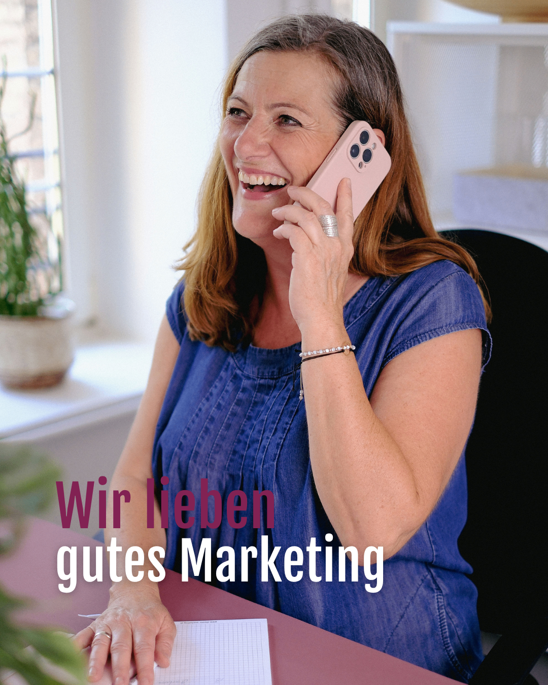 A woman in a blue shirt is sitting at a desk and talking on her pink phone. There is a plant on the windowsill behind her. Text overlay reads Wir lieben gutes Marketing.