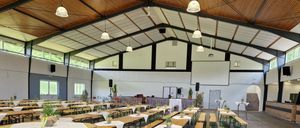 Ein Innenbankettsaal mit Holztischen und Bänken in Reihen angeordnet, dekoriert mit Pflanzen und ausgestattet mit Deckenleuchten und Lautsprechern.