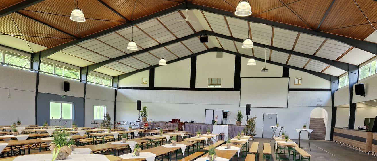 Ein Innenbankettsaal mit Holztischen und Bänken in Reihen angeordnet, dekoriert mit Pflanzen und ausgestattet mit Deckenleuchten und Lautsprechern.