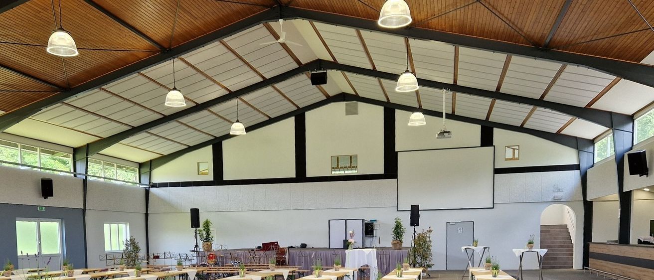Ein Innenbereich für das Essen mit Reihen von langen Holztischen und Bänken, jeweils mit dekorativen Pflanzenarrangements. Der Raum hat Holzbinder und Metallträger, mit einem Deckenventilator und hängenden Lampen.
