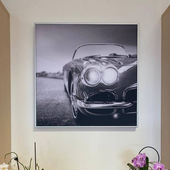 Ein schwarz-weißes Foto eines Oldtimers ist an einer weißen Wand über einem Holzregal montiert, das zwei weiße Vasen mit violetten und weißen Orchideen enthält.