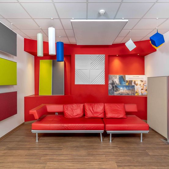 Ein moderner Loungebereich mit einem roten Ledersofa, farbenfrohen Hängelampen und einer abstrakten Wanddekoration. Der Raum hat Holzböden und eine weiße Decke.