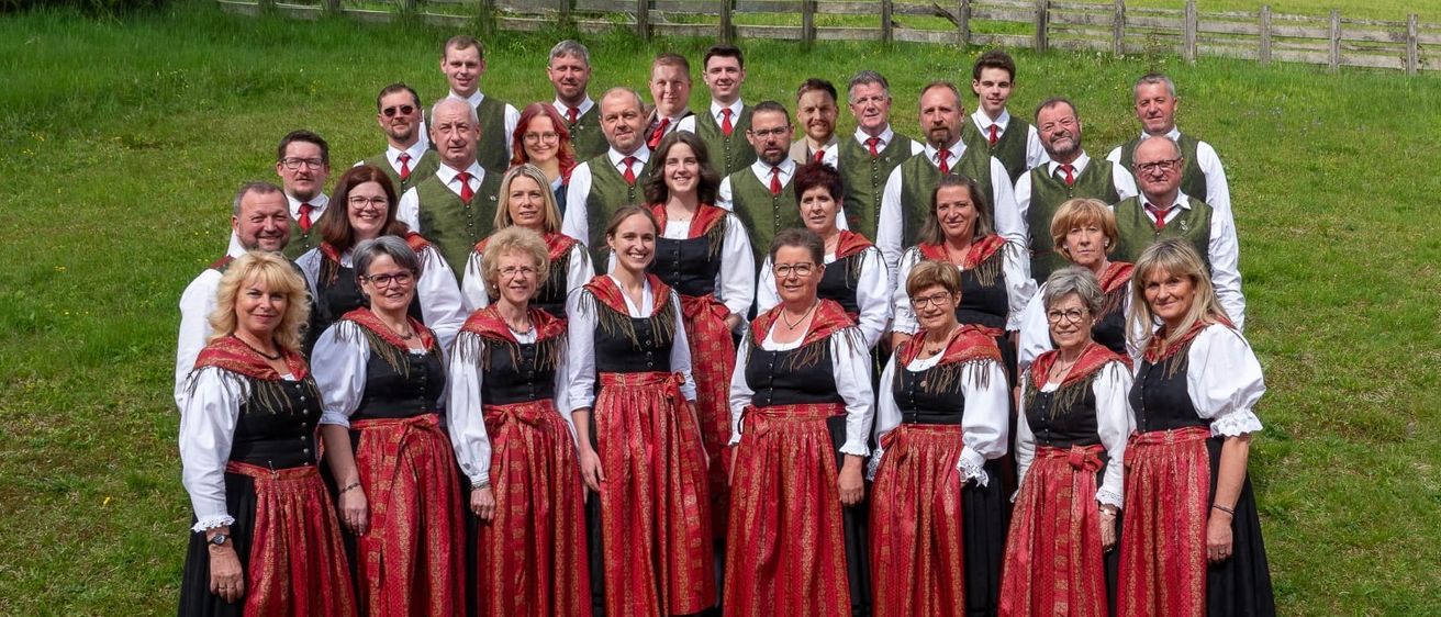 Eine Gruppe von Menschen in traditioneller Kleidung steht auf einer Rasenfläche, mit einem Holzzaun im Hintergrund. Einige tragen rote Krawatten, andere grüne Westen. Sie alle scheinen zu lächeln.