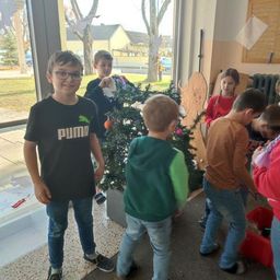 Eine Gruppe von Kindern schmückt einen Weihnachtsbaum in einem Flur. Der Junge im schwarzen Puma-Shirt steht links.
