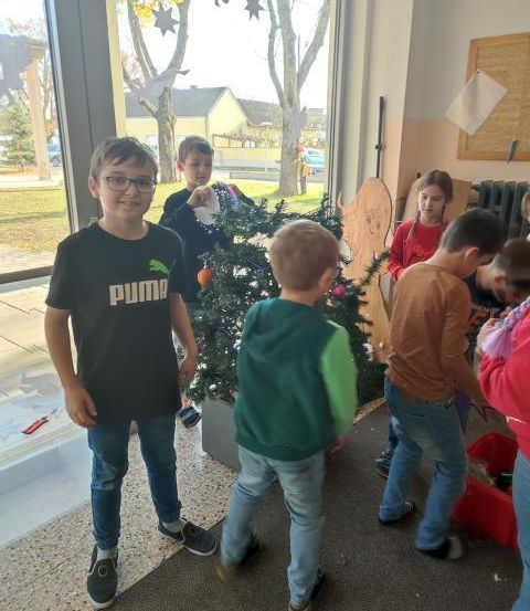 Eine Gruppe von Kindern schmückt einen Weihnachtsbaum in einem Flur. Der Junge im schwarzen Puma-Shirt steht links.