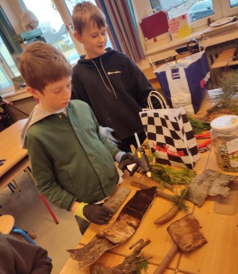 Zwei Jungen arbeiten mit Werkzeugen auf einem Tisch in einem Klassenzimmer, umgeben von verschiedenen Gegenständen wie einem Beutel und einem Glas.