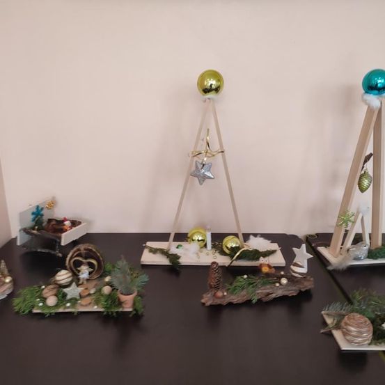 Ein Tisch zeigt eine Sammlung von Weihnachtsdekorationen, darunter verschiedene Ornamente, eine Kerze, ein Stern und eine Miniatur-Krippenszene in einer Schachtel. Es gibt drei dreieckige Ständer mit farbenfrohen Ornamenten obenauf.
