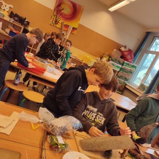 Schüler arbeiten in einem Klassenzimmer an einem Kunstprojekt. Ein Schüler hält ein großes Stück Holz, während ein anderer es genau betrachtet. Das Klassenzimmer ist mit verschiedenen Materialien und Werkzeugen gefüllt.