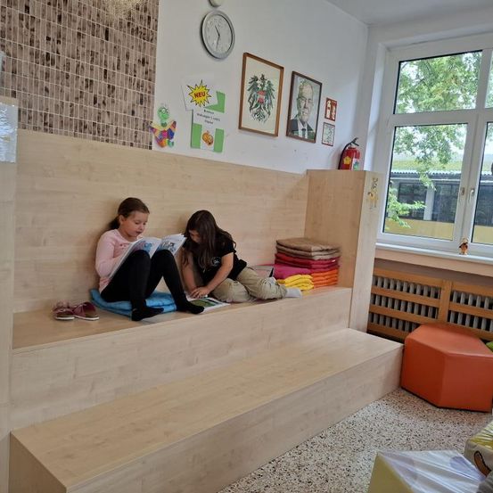Zwei junge Mädchen sitzen auf einer Holzbank in einem Raum und lesen Bücher. Ein Feuerlöscher steht an der Wand neben dem Fenster.