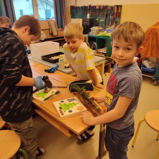 Drei Jungen arbeiten an Weihnachtsbasteleien an einem Tisch. Ein Junge lächelt, während die anderen sich auf ihre Projekte konzentrieren. Hinter ihnen ist ein Klassenzimmer mit Regalen und einem Fenster zu sehen.
