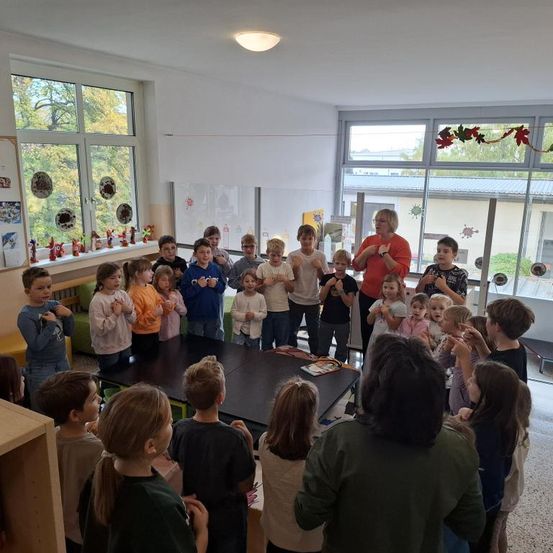 Eine Gruppe von Kindern steht in einem Klassenzimmer und sieht einer Frau zu, die eine Lektion zu führen scheint. Der Raum hat große Fenster und Dekorationen.