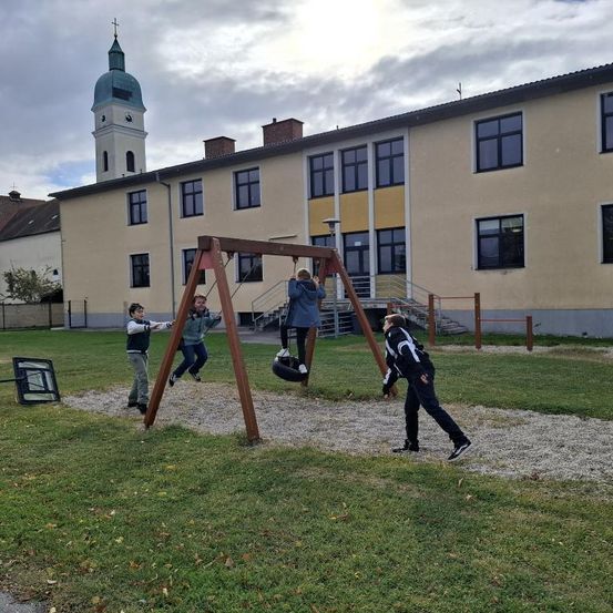 Kinder spielen auf einem Spielplatz im Hof eines Schulgebäudes, ein weiteres Kind läuft in der Nähe.