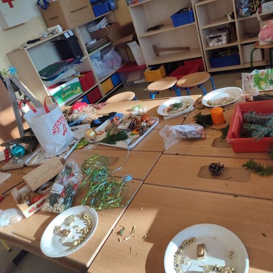 Ein Klassenzimmer mit einem Tisch voller Bastelmaterialien, einschließlich Pappteller, Bänder, Ornamente und Tannenzapfen. Der Raum ist auch mit Regalen und Lagerboxen gefüllt.