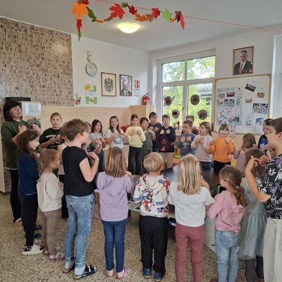 Eine Gruppe von Kindern und einer Frau stehen in einem Raum. Sie scheinen einen Tanz zu machen. An der Wand hängen Poster.