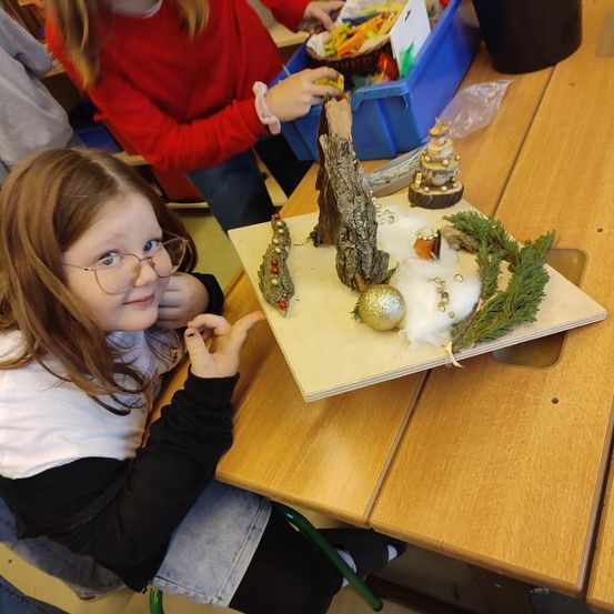 Ein junges Mädchen mit Brille sitzt an einem Tisch mit einem Weihnachtsbastelprojekt. Sie lächelt und zeigt. Ein Kind in einem roten Shirt steht hinter ihr und greift in eine blaue Kiste.
