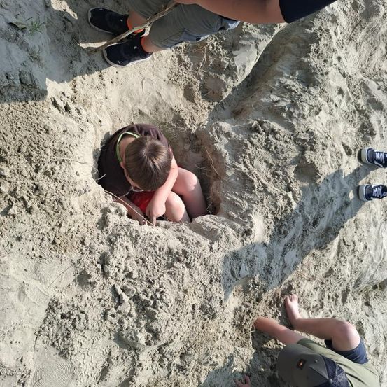 Ein Junge gräbt ein Loch im Sand, während zwei andere zusehen und einer einen Stock hält.