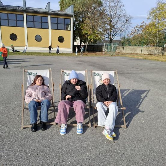 Drei Frauen sitzen auf Klappstühlen in einem Parkplatz und lächeln für ein Foto. Hinter ihnen steht ein Gebäude mit runden Fenstern, und mehrere Personen gehen in der Ferne.
