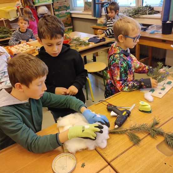 Mehrere junge Jungen sind in einem Klassenzimmer und beschäftigen sich mit einem Kunstprojekt. Sie tragen Handschuhe und haben Materialien wie Baumwolle und Tannenzweige auf ihren Tischen. Einige der Kinder benutzen Werkzeuge.