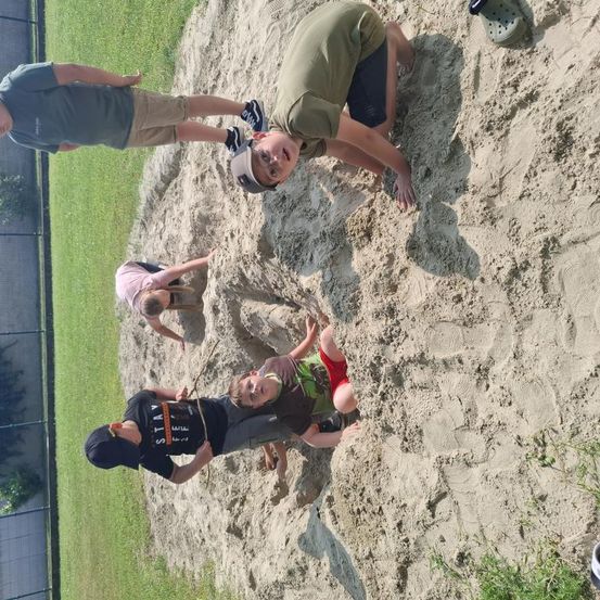 Fünf Kinder spielen in einem Sandkasten, wobei eines kniet und ein anderes auf dem Sand liegt. Einer hält einen Stock, und Fußabdrücke sind sichtbar. Ein Zaun und Gras umgeben das Gebiet.