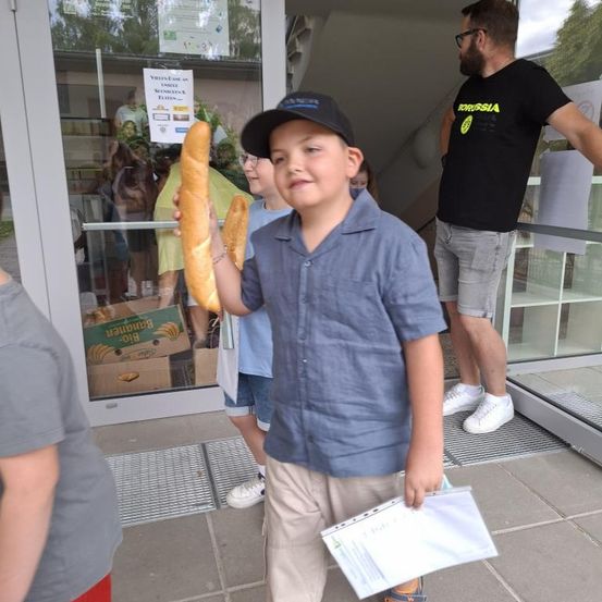 Ein Junge hält zwei Baguettes und lächelt vor einem Ladeneingang. Ein Mann in einem schwarzen T-Shirt steht hinter ihm, während Menschen sich im Laden bewegen.