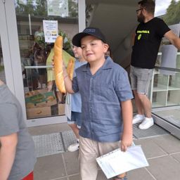 Ein Junge hält zwei Baguettes und lächelt vor einem Ladeneingang. Ein Mann in einem schwarzen T-Shirt steht hinter ihm, während Menschen sich im Laden bewegen.