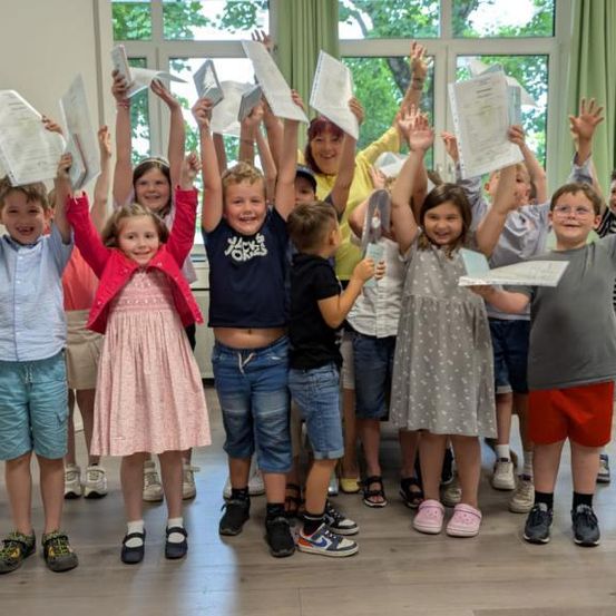 Eine Gruppe von Kindern in einem Klassenzimmer hält Urkunden mit glücklichen Gesichtsausdrücken. Sie stehen in einer Reihe, heben ihre Hände und lächeln zur Kamera.
