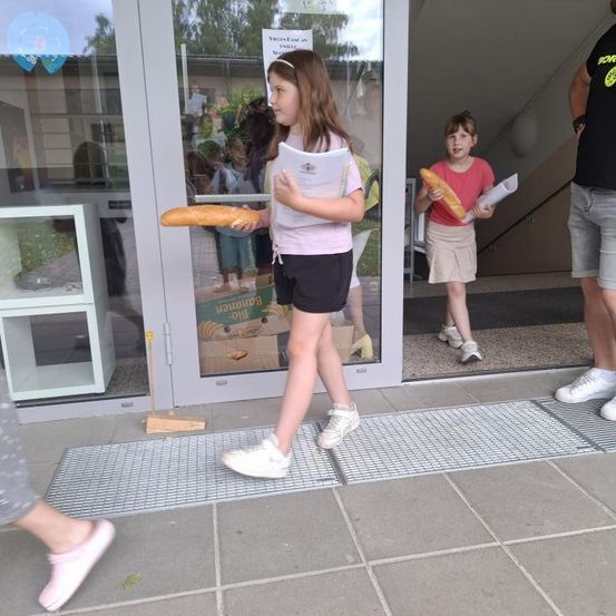 Zwei junge Mädchen tragen Baguettes und Papiere und verlassen ein Gebäude mit einer Glastür. Ein Mann steht in der Nähe und hält ein Blatt Papier.