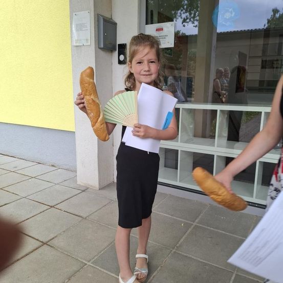 Ein kleines Mädchen steht draußen und hält ein Baguette und einen Fächer, mit einem Papier in der Hand und einer Person, die ein weiteres Baguette hält, hinter ihr.