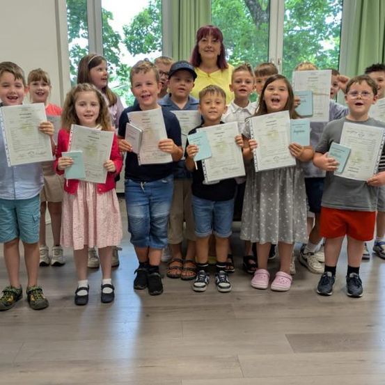 Eine Gruppe von Kindern und ihrer Lehrerin posiert für ein Foto, alle halten Urkunden in den Händen. Sie stehen in einem Raum mit Holzboden.