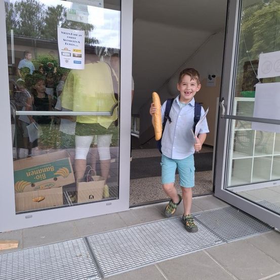 Ein Junge mit Rucksack tritt aus einer Glastür. Er hält eine Baguette. Leute stehen draußen, und eine Person in einem gelben Shirt ist durch das Glas sichtbar.
