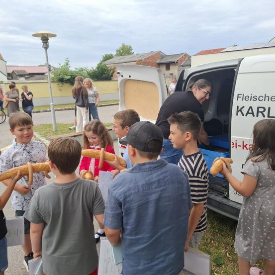 Kinder versammeln sich um einen Lieferwagen mit der Aufschrift 'Fleische Karl', halten Brot und interagieren mit einem lächelnden Erwachsenen, der einen Tablett mit Brot hält.