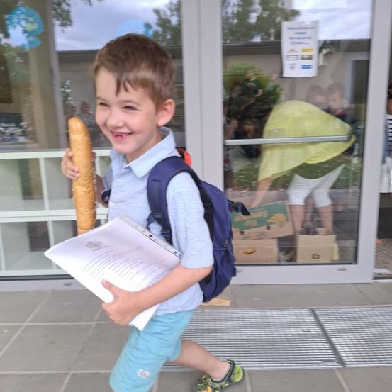 Ein junger Junge in einem blauen Hemd, Shorts und Turnschuhen verlässt ein Gebäude und hält einen langen Baguette und ein Papier in der Hand. Ein Rucksack ist auf seinem Rücken. Eine Frau in Gelb ist drinnen.