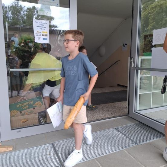 Ein Junge hält eine Baguette und geht auf den Eingang eines Gebäudes zu. Die Glastür spiegelt Menschen und ein Schild wider.