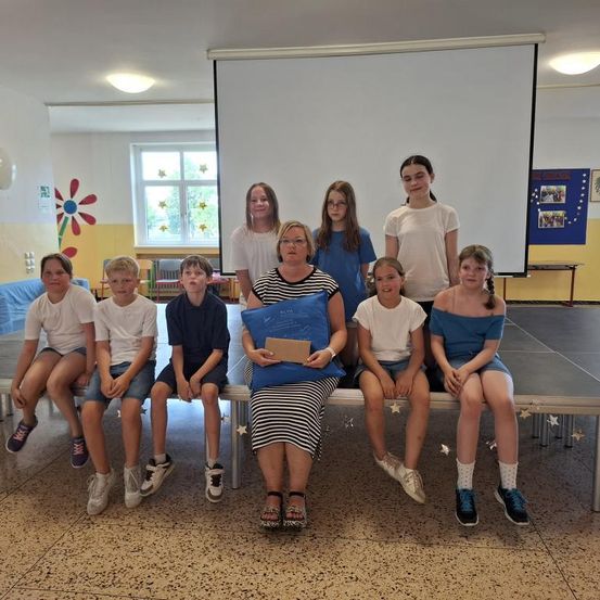 Eine Gruppe von Schülern und einer Lehrerin sitzen auf einer Bank in einem Klassenzimmer und lächeln für ein Foto. Die Lehrerin hält ein Zertifikat und eine Mappe.