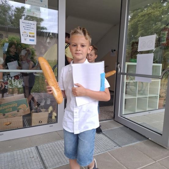 Ein junger Junge steht vor einem Gebäude, hält einen langen Baguette und eine Mappe. Hinter ihm sind mehrere Personen durch die Glastür sichtbar.
