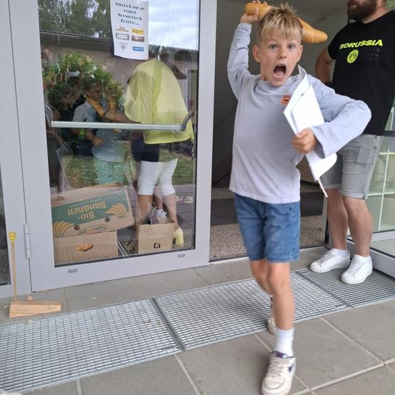 Ein Junge hält eine Baguette auf dem Kopf und geht auf eine Glastür zu, hinter ihm steht ein Mann mit einem Borussia-Shirt. Menschen warten draußen vor der Tür in einer Schlange.