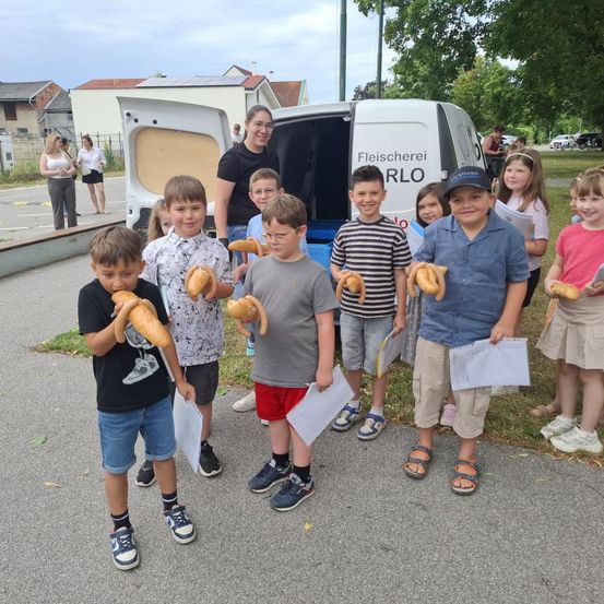 Eine Gruppe von Kindern, die Brot und Papiere vor einem Van mit dem Wort Fleischerei halten. Dahinter steht eine Frau, und einige Leute sind auf dem Bürgersteig. Im Hintergrund befinden sich Häuser, Autos und Bäume.