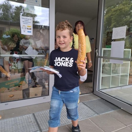 Ein junger Junge steht draußen und hält eine Baguette in der Hand, er trägt ein Jack & Jones-Shirt. Hinter ihm steht eine Frau in der Nähe einer Tür mit einem Schild. Im Inneren versammeln sich Leute in der Nähe eines Tisches mit Brotkisten.