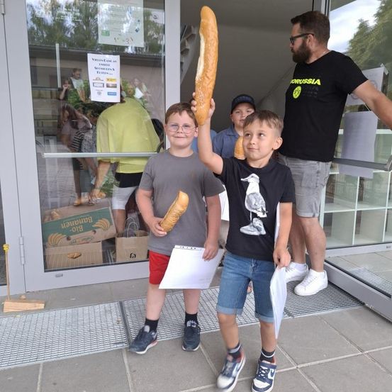 Zwei junge Jungen halten große Baguettes vor einem Geschäft. Ein Mann steht hinter ihnen und hält eine Mappe. Einige Personen sind durch die Glastür sichtbar.