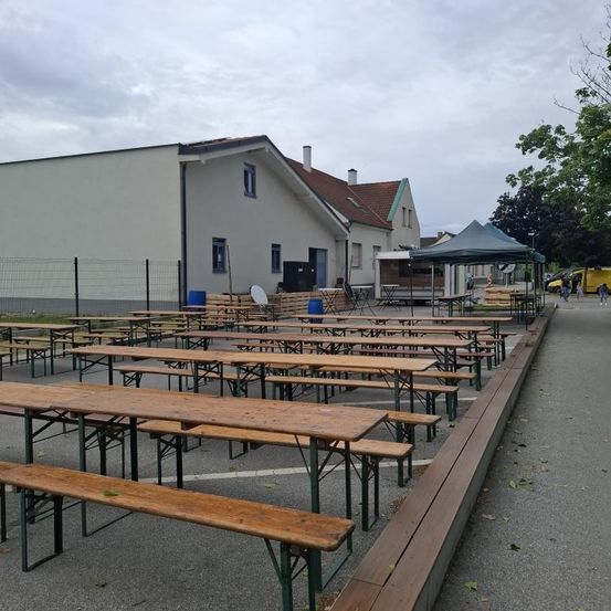 Eine Außensitzfläche mit Holztischen und Bänken. Dahinter ein Gebäude mit rotem Dach. Ein Zelt ist aufgebaut, und es gibt Fahrzeuge und Personen in der Nähe.