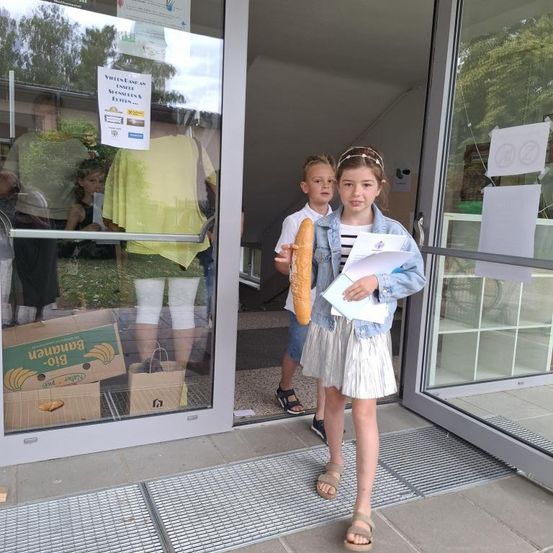 Zwei Kinder stehen vor einem Ladeneingang, eines hält ein Baguette und das andere einen Zettel. Sie sind im Glastür reflektiert. Es gibt Bananen und andere Gegenstände im Laden.