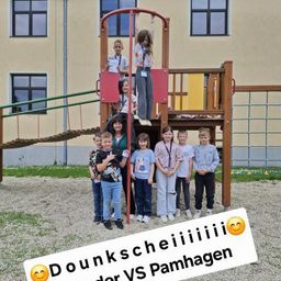 Eine Gruppe von Kindern steht in einem Spielplatz mit einer Frau. Der Spielplatz hat eine Rutsche und ein Klettergerüst. Das Bild hat einen Text-Overlay mit der Aufschrift 'Dounks VS Pamhagen'.