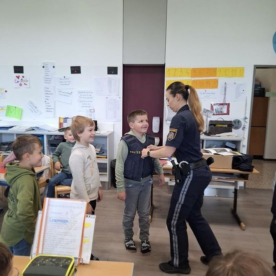 Zwei Jungen stehen lächelnd vor einer Polizistin im Klassenzimmer. Die Frau hält die Hand eines Jungen, und mehrere Kinder sitzen an Tischen im Hintergrund. Papiere und Poster sind an den Wänden befestigt.