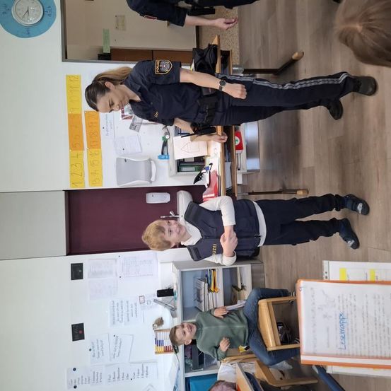 Eine Polizistin steht in einem Klassenzimmer mit Schülern, einer hält ein Abzeichen, ein anderer sitzt. Papiere, eine Uhr und Bücher sind an der Wand.