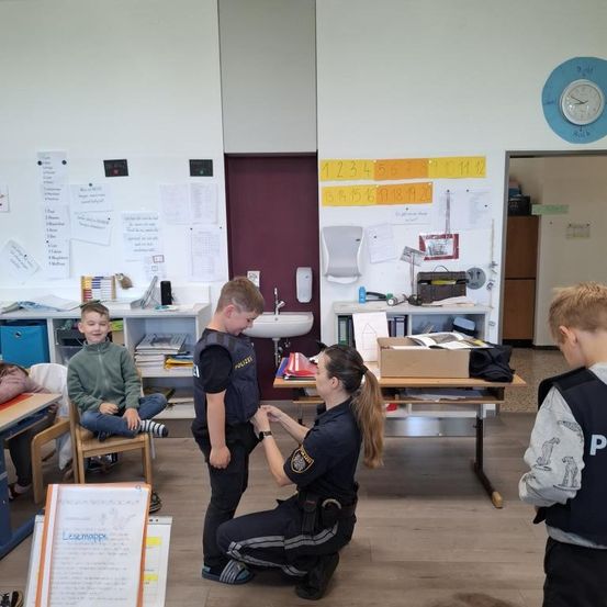 In einem Klassenzimmer interagiert eine Polizistin mit zwei Jungen, einer kniet und der andere steht. Ein weiterer Junge steht in der Nähe. Der Raum hat Schreibtische, ein Waschbecken und Bulletin-Boards.