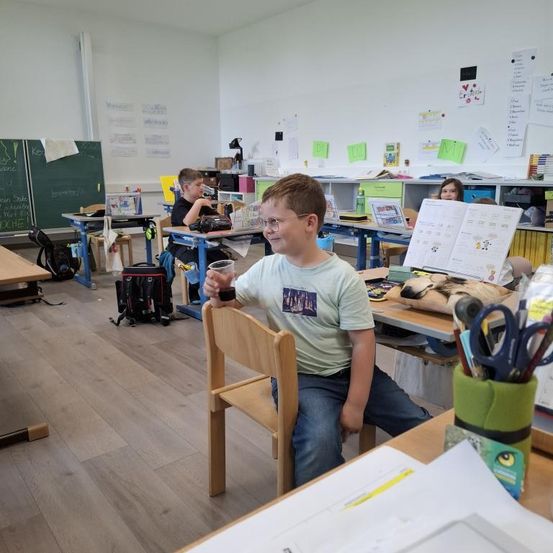 Ein Junge in einem Klassenzimmer hält einen Becher und lächelt, während er auf einem Stuhl sitzt. Hinter ihm arbeiten andere Schüler an Schreibtischen. Das Klassenzimmer hat einen Holzboden, Schreibtische, Stühle und verschiedene Poster an der Wand.