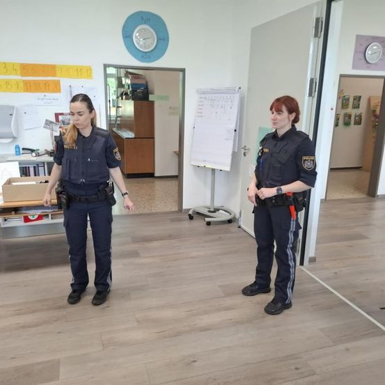 Zwei Polizeibeamte stehen in einem Klassenzimmer. Einer schaut etwas an, während der andere nach rechts schaut. Vor ihnen steht eine Tafel.