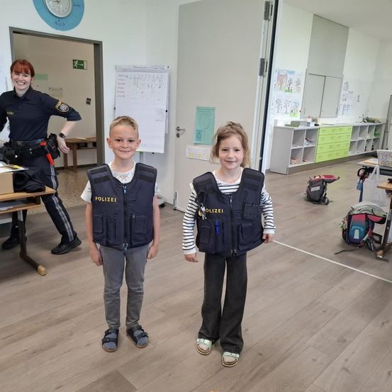 Zwei Kinder in Polizeiwesten stehen in einem Klassenzimmer mit einer lächelnden Polizeibeamtin im Hintergrund.