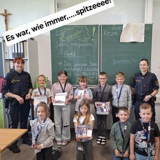 Eine Gruppe von Kindern steht lächelnd in einem Klassenzimmer. Sie halten Urkunden in den Händen und sind von zwei Polizeibeamten und einer Lehrerin umgeben. Tafeln mit Schriftzügen im Hintergrund.
