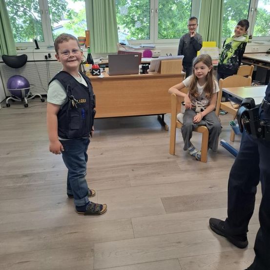 Mehrere Kinder sind in einem Klassenzimmer. Ein Junge steht und trägt eine Polizeiweste, während ein Mädchen auf einem Stuhl sitzt. Dahinter stehen zwei andere Kinder.